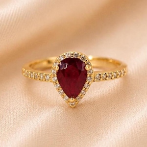 Könnte beinhalten: Ein Goldring mit einem birnenförmigen, tiefroten Rubin in der Mitte, der von einem Kranz aus kleinen, klaren weißen Steinen umgeben ist. Die Ringschiene ist zudem mit einer Reihe kleiner, klarer weißer Steine an beiden Seiten besetzt.