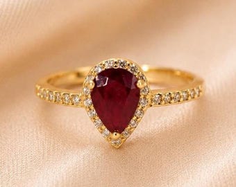 May include: A gold ring featuring a pear-shaped deep red ruby centre stone surrounded by a halo of small white round-cut stones. The band is also set with a row of small white round-cut stones extending along both sides of the central setting.