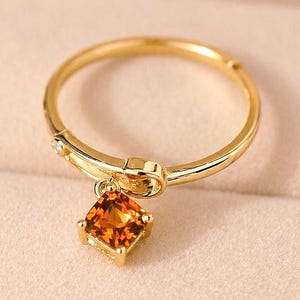 Könnte beinhalten: Ein goldener Ring mit einem quadratischen, orangefarbenen Edelsteinanhänger. Der Ring hat ein zartes Design mit einem kleinen Akzentstein am Band. Der Edelstein hat eine warme, bernsteinfarbene Farbe, und der Ring ist vor einem weichen, neutralen Hintergrund platziert.