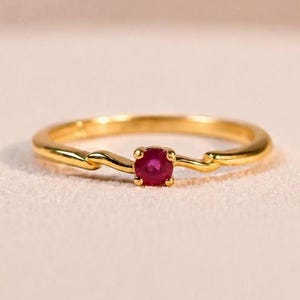 Könnte beinhalten: Ein goldener Ring mit einem einzelnen, runden, tiefroten Rubin, der in einer Vier-Krappen-Fassung sitzt. Die Ringschiene hat eine glatte, polierte Oberfläche und ein dezentes, wellenförmiges Design, das zum zentralen Stein führt.