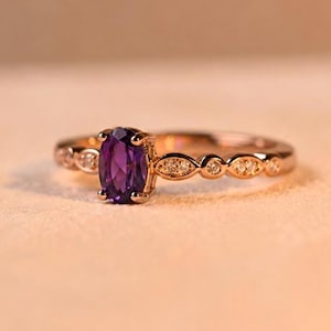 Op de afbeelding: Een roségouden ring met een centrale ovaal geslepen paarse amethist in een zetting met vier pootjes. De ringband is versierd met afwisselende markiesvormige en ronde accenten, elk ingelegd met kleine, heldere, sprankelende witte steentjes.