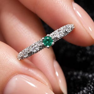 Puede incluir: Un anillo de tono plateado con una piedra preciosa verde de corte redondo en el centro, flanqueada por piedras más pequeñas y claras engastadas en una banda texturizada. El anillo metálico tiene un acabado granulado tipo pavé que se extiende por la superficie, resaltando el contraste entre la piedra central verde y los acentos claros.