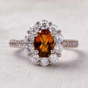 Peut inclure: Une bague avec une pierre centrale ovale de couleur ambre entourée d'un halo de douze pierres claires rondes. L'anneau est métallique et comporte une rangée de petites pierres claires incrustées le long des côtés de la tige.