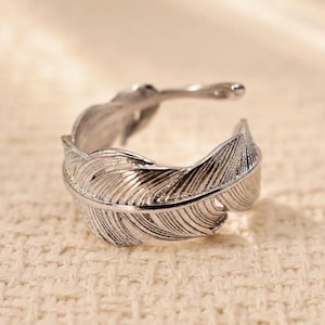 Könnte beinhalten: Ein silberner Federring. Der Ring hat ein detailliertes Federdesign, mit Linien, die in das Metall geätzt sind, um die Textur einer echten Feder nachzuahmen. Der Ring ist so konzipiert, dass er sich um den Finger wickelt.