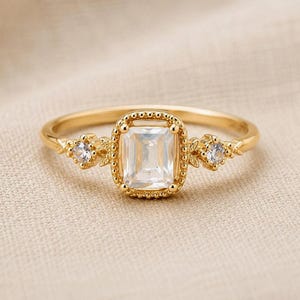 Pode incluir: Um anel de ouro com uma pedra central transparente de corte esmeralda rodeada por uma borda de contas. A banda inclui duas pedras redondas transparentes menores em cada lado, ladeadas por pequenos detalhes metálicos em forma de folha.