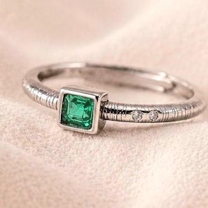 Könnte beinhalten: Ein silberfarbener Ring mit einem quadratisch geschliffenen grünen Edelstein in einer Fassung. Die Ringschiene hat eine texturierte, gerippte Oberfläche und enthält zwei kleine, klare runde Steine an einer Seite des Rings.