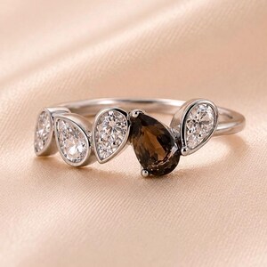 Könnte beinhalten: Ein silberfarbener Ring mit einer Anordnung von fünf birnenförmigen Steinen. Das Design umfasst vier klare, facettierte weiße Steine und einen zentralen, dunkelbraunen, birnenförmigen Stein, die alle in eine glatte, polierte Metallschiene gefasst sind.