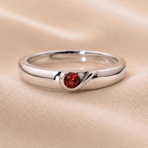 Peut inclure: Une bague en métal argenté poli dotée d'une unique petite pierre précieuse grenat rouge foncé, taille ronde, sertie dans un détail de canal incurvé et encastré sur la surface avant de l'anneau, créant un profil circulaire lisse et continu.