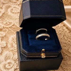 May include: Two metallic rings with a smooth, polished finish are displayed inside a dark navy blue, octagonal-shaped jewellery box. The box features a gold-toned latch with a small clear stone and a deep blue velvet interior lining.