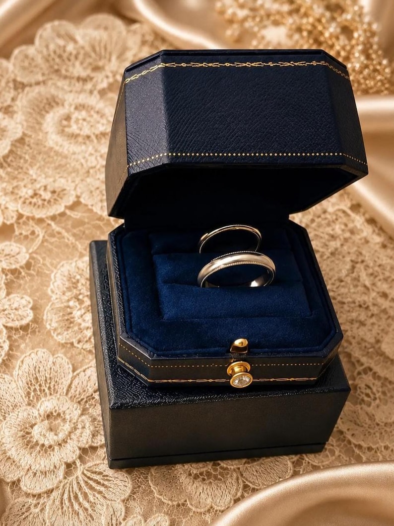 May include: Two metallic rings with a smooth, polished finish are displayed inside a dark navy blue, octagonal-shaped jewellery box. The box features a gold-toned latch with a small clear stone and a deep blue velvet interior lining.