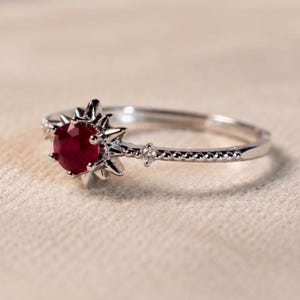 K&ouml;nnte beinhalten: Ein silberfarbener Ring mit einem runden, tiefroten Edelstein in einer sternf&ouml;rmigen Krappenfassung. Die Ringschiene hat eine feine Perlenstruktur und enth&auml;lt einen kleinen, klaren Akzentstein auf jeder Seite des zentralen Sternmotivs.