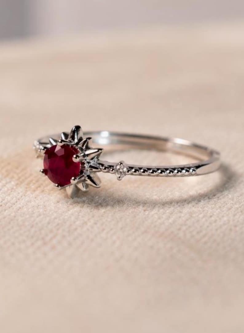 K&ouml;nnte beinhalten: Ein silberfarbener Ring mit einem runden, tiefroten Edelstein in einer sternf&ouml;rmigen Krappenfassung. Die Ringschiene hat eine feine Perlenstruktur und enth&auml;lt einen kleinen, klaren Akzentstein auf jeder Seite des zentralen Sternmotivs.