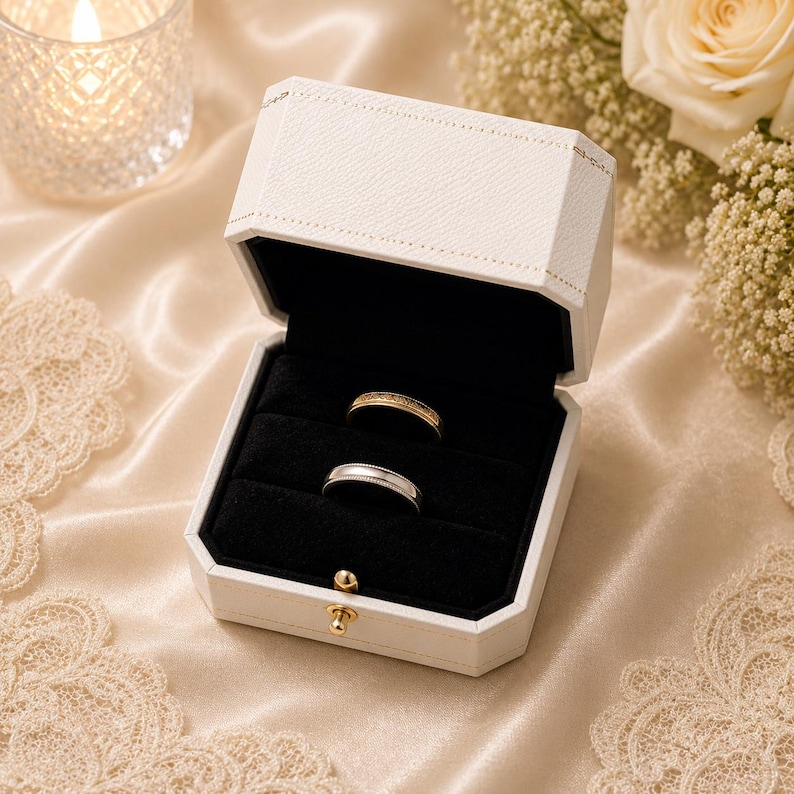 May include: An open white textured ring box with a black velvet interior containing two rings. One ring is gold with a textured surface, and the other is a smooth silver-toned metal band. The box features a small gold-coloured metal latch closure.