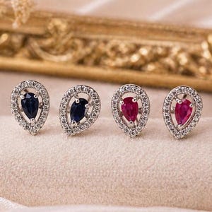 Puede incluir: Cuatro pendientes de botón en forma de lágrima dispuestos en fila. Dos pendientes tienen piedras azul oscuro en talla pera y dos tienen piedras rojo vibrante en talla pera. Cada piedra está rodeada por un halo de pequeñas piedras blancas.