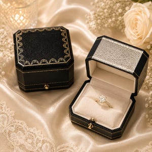 May include: A gold ring with a round-cut centre stone and a pav&eacute; band sits inside an open black octagonal jewellery box with a cream velvet interior. A closed matching black box with gold decorative trim sits beside it on a satin fabric surface.