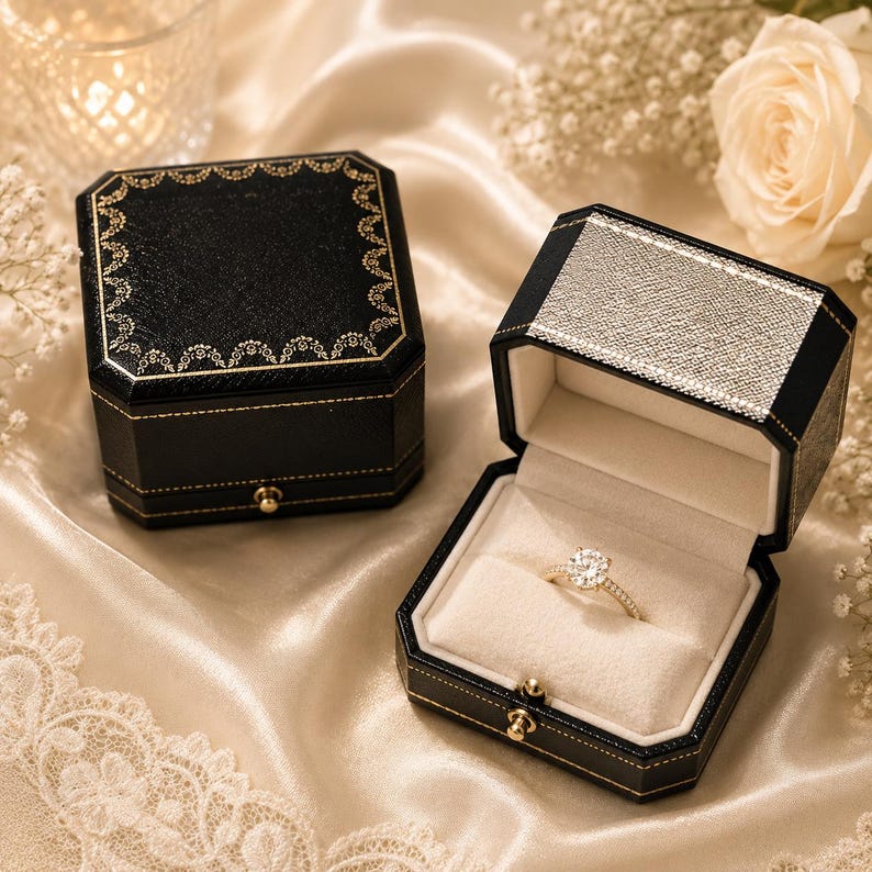 May include: A gold ring with a round-cut centre stone and a pav&eacute; band sits inside an open black octagonal jewellery box with a cream velvet interior. A closed matching black box with gold decorative trim sits beside it on a satin fabric surface.