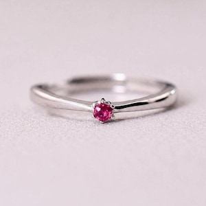 Può includere: Un anello color argento con una singola piccola pietra preziosa rotonda di colore rosa vivace, incastonata al centro con griffe. La fascia è liscia e lucida, con un leggero restringimento verso la pietra per un design pulito.