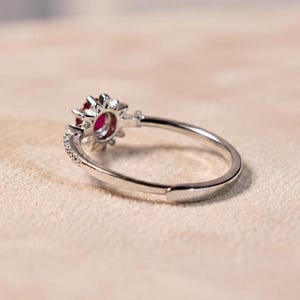 K&ouml;nnte beinhalten: Ein silberfarbener Ring mit einem zentralen runden roten Edelstein, der von einem Kranz aus kleinen klaren Steinen umgeben ist. Die Ringschiene ist d&uuml;nn und metallisch mit einer sichtbaren &Ouml;ffnung an der R&uuml;ckseite f&uuml;r ein verstellbares Design.