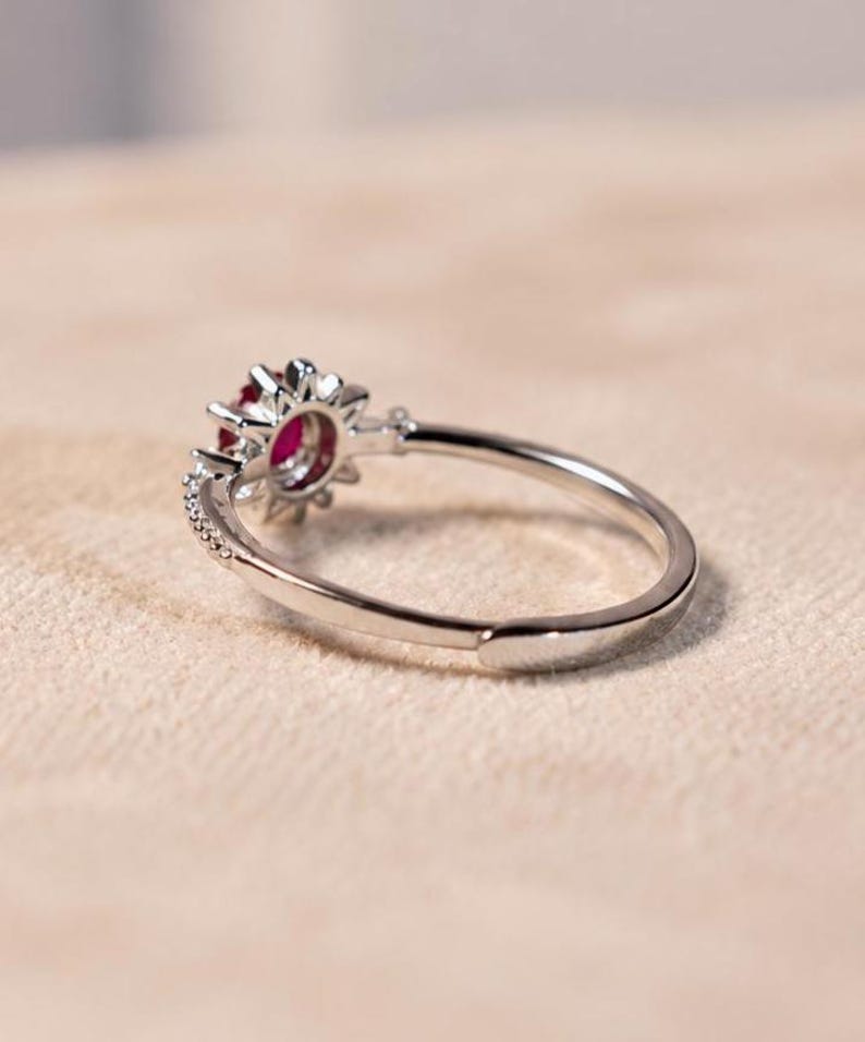 K&ouml;nnte beinhalten: Ein silberfarbener Ring mit einem zentralen runden roten Edelstein, der von einem Kranz aus kleinen klaren Steinen umgeben ist. Die Ringschiene ist d&uuml;nn und metallisch mit einer sichtbaren &Ouml;ffnung an der R&uuml;ckseite f&uuml;r ein verstellbares Design.