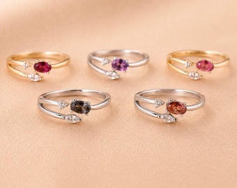 Puede incluir: Cinco anillos tipo bypass con piedras preciosas de corte ovalado y piedras claras de corte marquesa. Dos anillos son dorados con piedras rosas, uno es plateado con una piedra morada, uno es plateado con una piedra gris oscuro y uno es plateado con una piedra marrón rojiza.