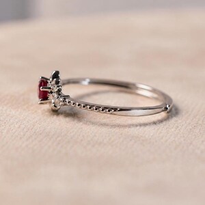 K&ouml;nnte beinhalten: Ein silberfarbener Ring mit einem kleinen, runden, tiefroten Edelstein in der Mitte. Die Ringschiene hat eine zarte, perlenartige Textur an den Seiten, die zu der Krappenfassung f&uuml;hrt, welche den roten Stein sicher an seinem Platz h&auml;lt.