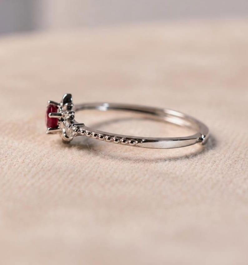 K&ouml;nnte beinhalten: Ein silberfarbener Ring mit einem kleinen, runden, tiefroten Edelstein in der Mitte. Die Ringschiene hat eine zarte, perlenartige Textur an den Seiten, die zu der Krappenfassung f&uuml;hrt, welche den roten Stein sicher an seinem Platz h&auml;lt.