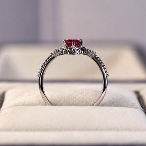 K&ouml;nnte beinhalten: Ein silberfarbener Ring mit einem runden, tiefroten Edelstein in der Mitte, der von einer Krappenfassung gehalten wird. Das Band ist an den Seiten mit kleinen, klaren, funkelnden Steinen besetzt und hat eine zarte, perlenartige Textur.