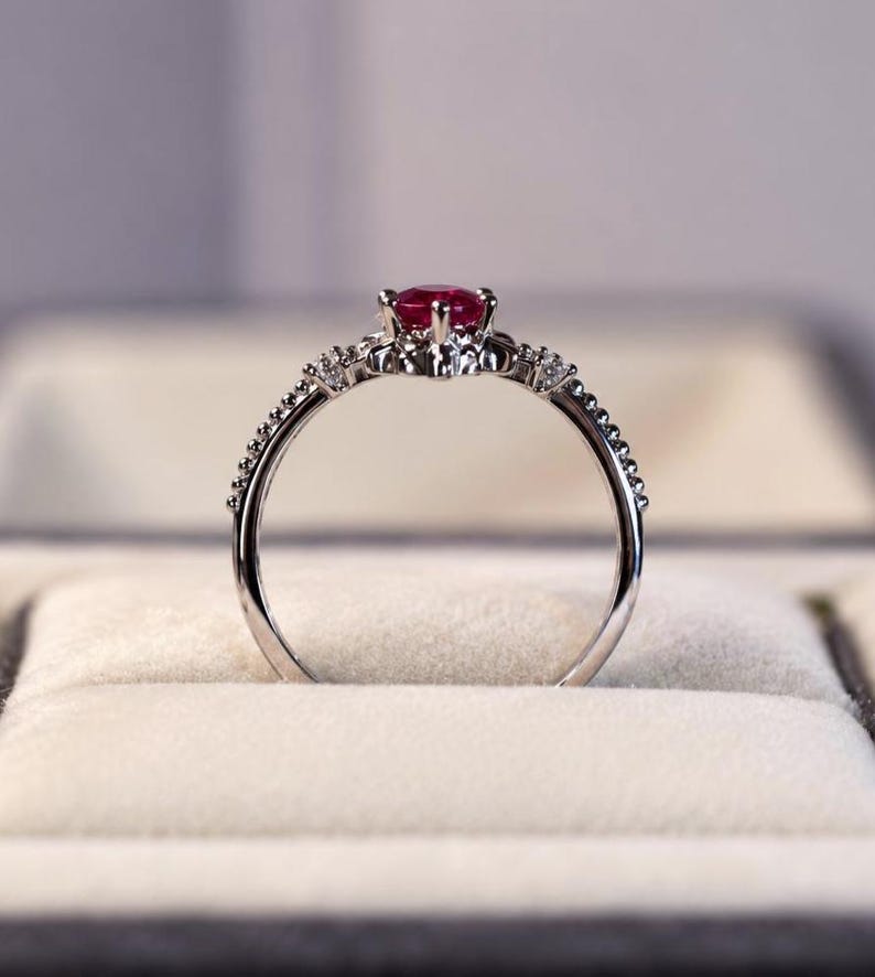 K&ouml;nnte beinhalten: Ein silberfarbener Ring mit einem runden, tiefroten Edelstein in der Mitte, der von einer Krappenfassung gehalten wird. Das Band ist an den Seiten mit kleinen, klaren, funkelnden Steinen besetzt und hat eine zarte, perlenartige Textur.