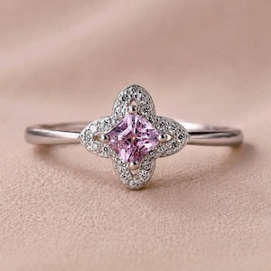 May include: A silver-toned ring featuring a central princess-cut light pink gemstone. The stone is set within a four-petaled floral-shaped frame encrusted with small clear white stones, mounted on a smooth, polished silver-toned metal band.