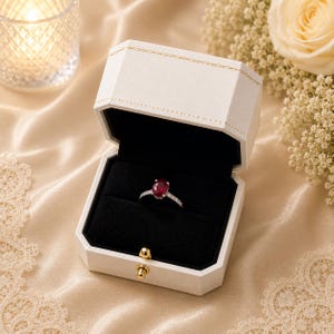 May include: A silver ring featuring a central oval-cut red ruby surrounded by small clear stones on the band, presented inside an open white hexagonal jewellery box with a black velvet interior and a gold-toned metal latch closure.