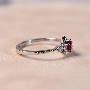 K&ouml;nnte beinhalten: Ein silberfarbener Ring mit einem zentralen, rund geschliffenen roten Edelstein, der von einer Gruppe kleiner, klarer, funkelnder Steine umgeben ist. Die Ringschiene hat eine feine, perlenartige Textur an den Seiten bis zur Fassung.