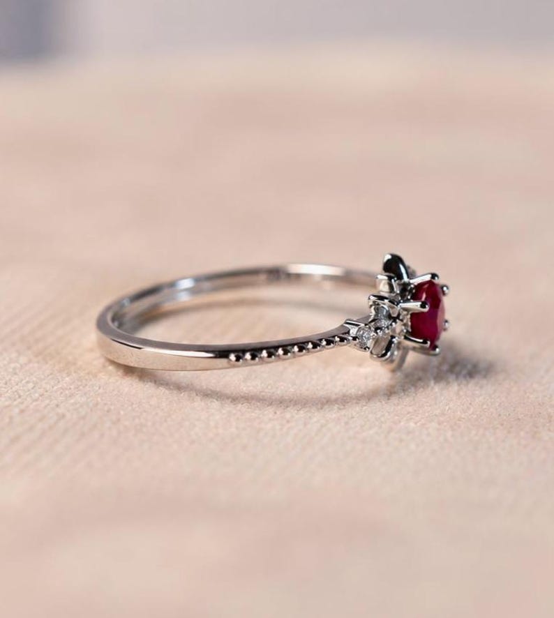 K&ouml;nnte beinhalten: Ein silberfarbener Ring mit einem zentralen, rund geschliffenen roten Edelstein, der von einer Gruppe kleiner, klarer, funkelnder Steine umgeben ist. Die Ringschiene hat eine feine, perlenartige Textur an den Seiten bis zur Fassung.