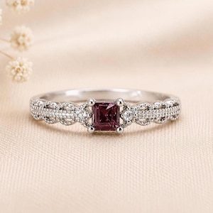 Könnte beinhalten: Ein silberfarbener Ring mit einem quadratisch geschliffenen violetten Edelstein in der Mitte. Die Ringschiene zeigt ein filigranes, texturiertes Muster mit kleinen, klaren Akzentsteinen, die die zentrale Fassung auf beiden Seiten flankieren.