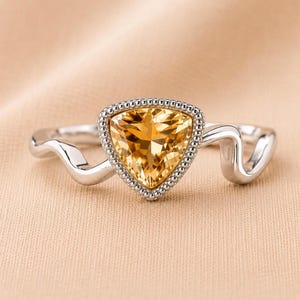 Peut inclure: Une bague argentée ornée d'une pierre précieuse citrine jaune triangulaire à facettes en son centre. La pierre est entourée d'une bordure métallique perlée, et l'anneau présente un design ondulé distinct qui se courbe le long du doigt.