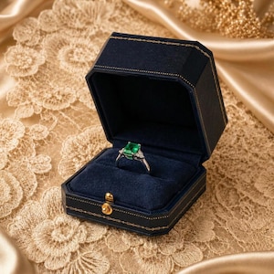 May include: A silver ring featuring a rectangular emerald-cut green gemstone flanked by small clear stones, presented inside an open navy blue hexagonal jewellery box with gold-tone trim and a dark blue velvet interior lining.