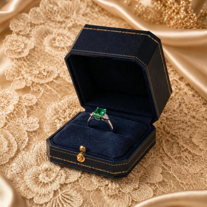 May include: A silver ring featuring a rectangular emerald-cut green gemstone flanked by small clear stones, presented inside an open navy blue hexagonal jewellery box with gold-tone trim and a dark blue velvet interior lining.
