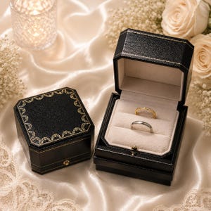 May include: Two black octagonal ring boxes with gold decorative trim. One box is closed, and the other is open, revealing a beige velvet interior holding a thin gold band and a silver-toned band. The boxes feature a textured exterior finish.