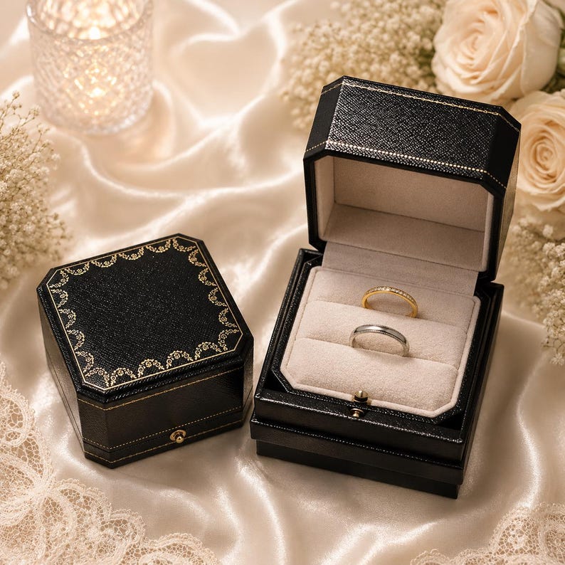 May include: Two black octagonal ring boxes with gold decorative trim. One box is closed, and the other is open, revealing a beige velvet interior holding a thin gold band and a silver-toned band. The boxes feature a textured exterior finish.