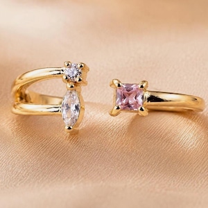 Könnte beinhalten: Ein goldfarbener offener Ring mit geteiltem Banddesign. Eine Seite zeigt einen runden, klaren Stein und einen klaren Stein im Marquise-Schliff. Die andere Seite weist einen einzelnen quadratischen, rosafarbenen Stein auf. Jeder Stein ist in einer Vier-Krappen-Fassung gefasst.