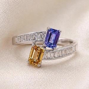 Puede incluir: Un anillo tipo bypass de metal plateado con dos piedras preciosas de corte esmeralda, una azul y otra amarilla, engastadas en el centro. La banda está decorada con una hilera de pequeñas piedras transparentes de corte baguette a cada lado.