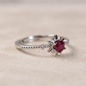 K&ouml;nnte beinhalten: Ein silberfarbener Ring mit einem zentralen, rund geschliffenen, tiefroten Rubin, der in eine sonnenstrahlenf&ouml;rmige Fassung eingefasst ist. Die Ringschiene ist mit kleinen Perldetails und einem winzigen klaren Stein verziert.