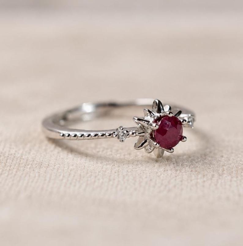 K&ouml;nnte beinhalten: Ein silberfarbener Ring mit einem zentralen, rund geschliffenen, tiefroten Rubin, der in eine sonnenstrahlenf&ouml;rmige Fassung eingefasst ist. Die Ringschiene ist mit kleinen Perldetails und einem winzigen klaren Stein verziert.