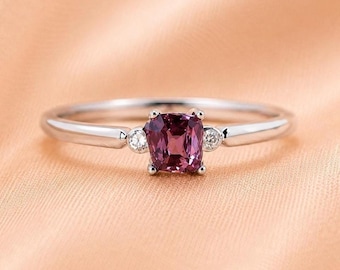 Puede incluir: Un anillo de tono plateado con una piedra preciosa central de color rosa intenso en talla cojín, flanqueada por dos pequeñas piedras redondas y transparentes. El aro es liso y pulido, con un engaste metálico sencillo para las piedras.