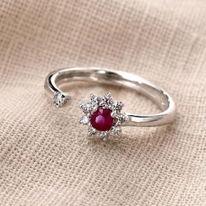 Può includere: Un anello aperto color argento con una pietra rossa rotonda centrale circondata da un alone di piccole pietre chiare. Un'estremità della fascia termina con una piccola pietra chiara, mentre l'altra mostra un gruppo di pietre a forma di fiore.