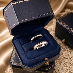 May include: Two metallic rings with a dual-tone finish resting inside an open, navy blue, octagonal jewellery box. The box features a textured exterior with gold-coloured decorative trim and a deep blue velvet interior lining for the rings.