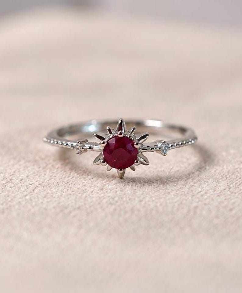 K&ouml;nnte beinhalten: Ein silberfarbener Ring mit einem zentralen, rund geschliffenen, tiefroten Edelstein in einer sternf&ouml;rmigen Fassung. Die Ringschiene ist auf beiden Seiten mit kleinen, klaren Steinen besetzt und weist eine feine Perlenstruktur auf.