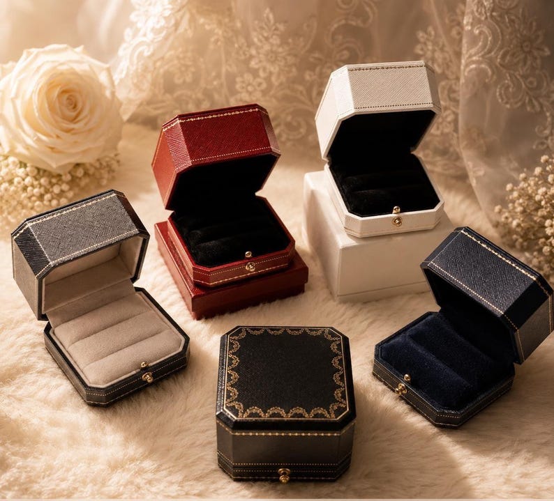 May include: A collection of five octagonal jewellery boxes in various colours including dark grey, deep red, white, black, and navy blue. Each box features a textured exterior, a gold-toned metal clasp, and a soft velvet interior lining for rings.