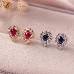 Può includere: Due paia di orecchini a lobo a forma di goccia. Un paio presenta montature color oro con pietre centrali rosso scuro circondate da cristalli chiari. Il secondo paio ha montature color argento con pietre centrali blu scuro e cristalli.