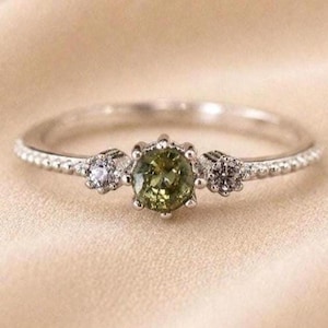 Könnte beinhalten: Ein silberfarbener Ring mit einem zentralen, rund geschliffenen olivgrünen Edelstein, der von zwei kleineren, klaren, rund geschliffenen Steinen flankiert wird. Die Ringschiene hat eine feine Perlenstruktur an den Seiten.