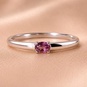 Könnte beinhalten: Ein Ring aus poliertem, silberfarbenem Metall mit einem einzelnen, kleinen, ovalen rosa Edelstein, der in einer Vier-Krappen-Fassung in der Mitte der Ringschiene sitzt. Die Schiene ist glatt, abgerundet und hat eine gleichmäßige Breite.